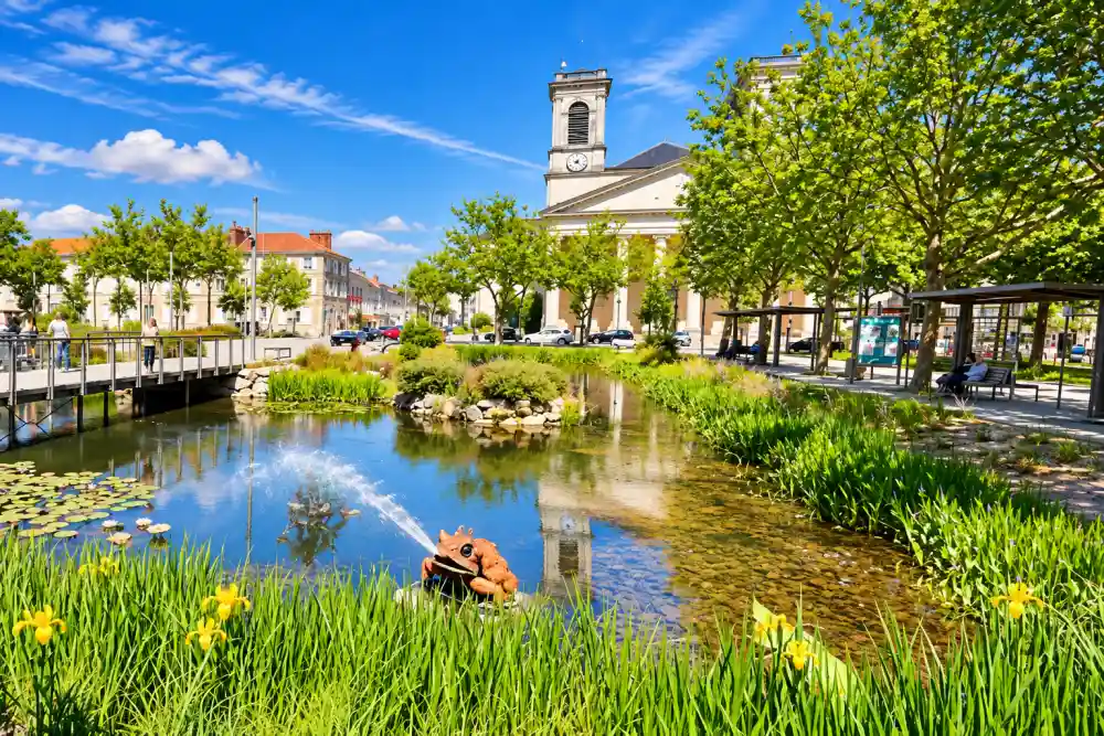 Investir à La Roche-sur-Yon : quels quartiers choisir pour un investissement immobilier rentable ? 2 étang bordé de nénuphars et de joncs, pont piétonnier à gauche, arbres alignés et bâtiment avec horloge en arrière-plan sous ciel bleu