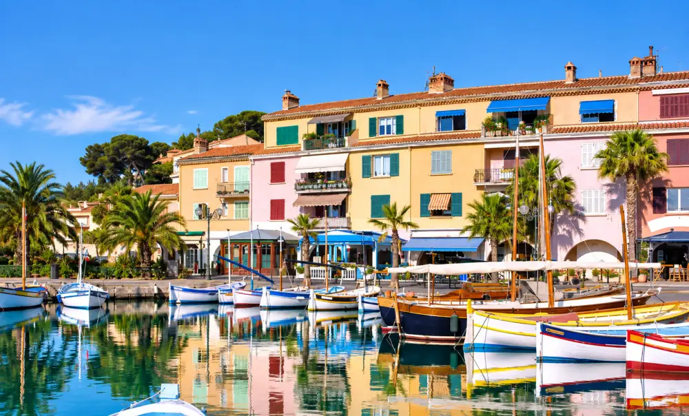 Vivre à Six-Fours-les-Plages en 2026 : immobilier, prix et qualité de vie 3 vue sur quai bordé de maisons pastel à volets verts et rouges, palmiers et bateaux colorés reflétés dans l’eau.